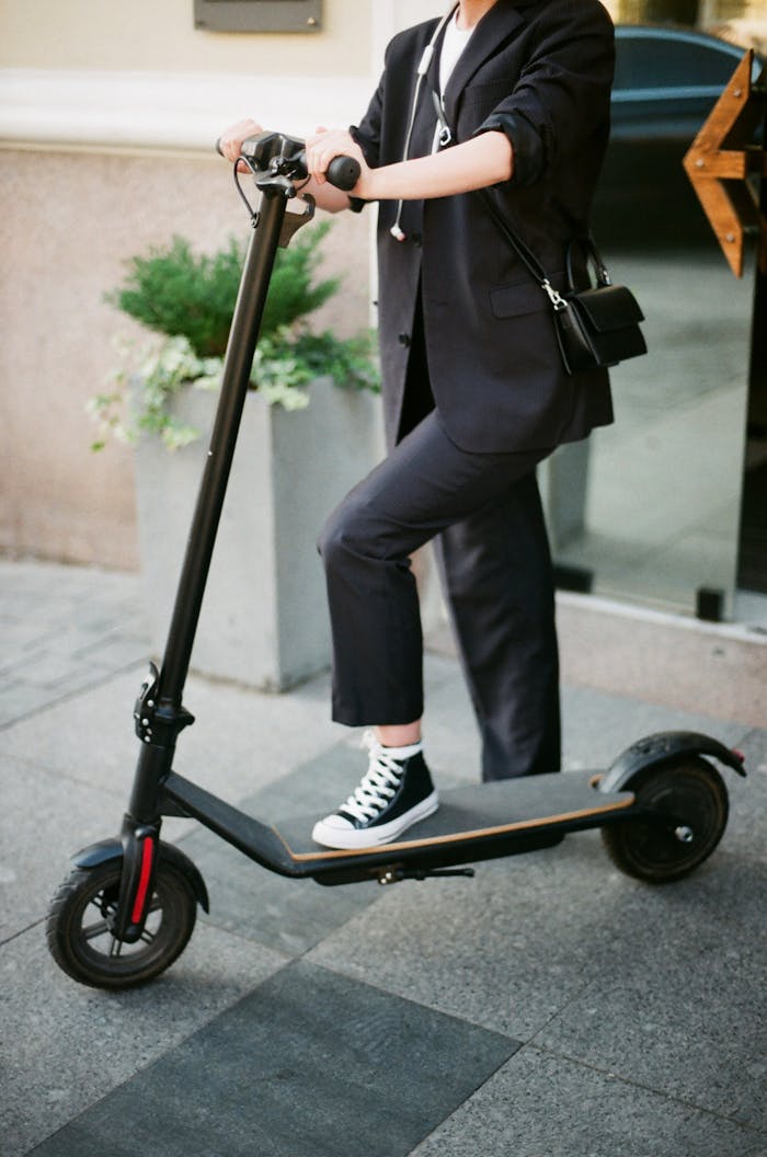 A fashionable woman in a suit rides an electric scooter on a city street.